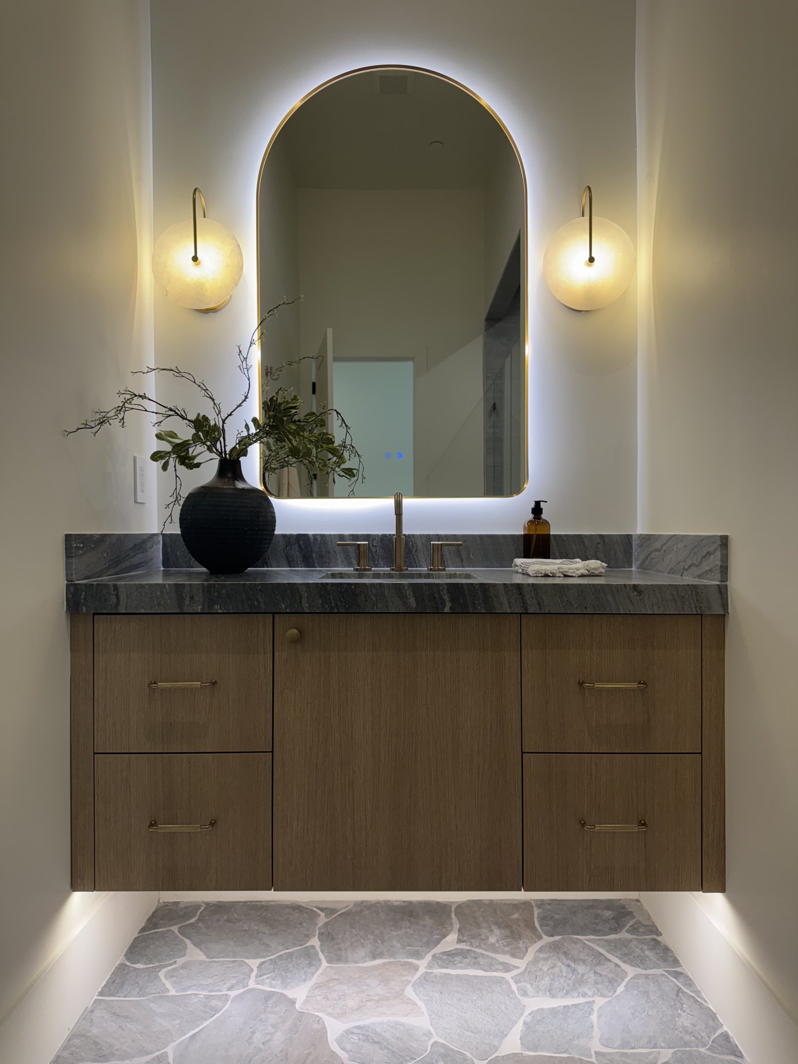 Chandler AZ guest bathroom with ash-toned floating vanity, dark veined stone vanity top, arched backlit mirror, brass fixtures, and flagstone-pattern porcelain floor