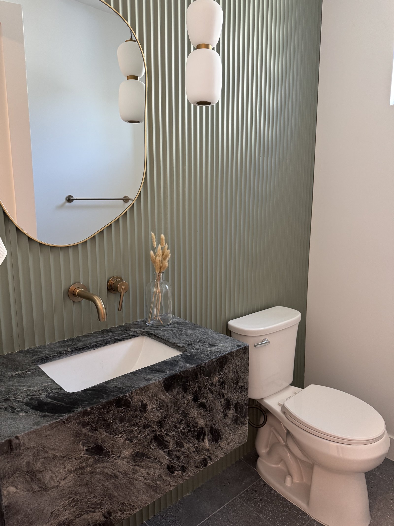 Chandler AZ powder room with sage-green fluted accent wall, brass-detailed round mirror, dark veined marble waterfall vanity, and twin brass pendant lights