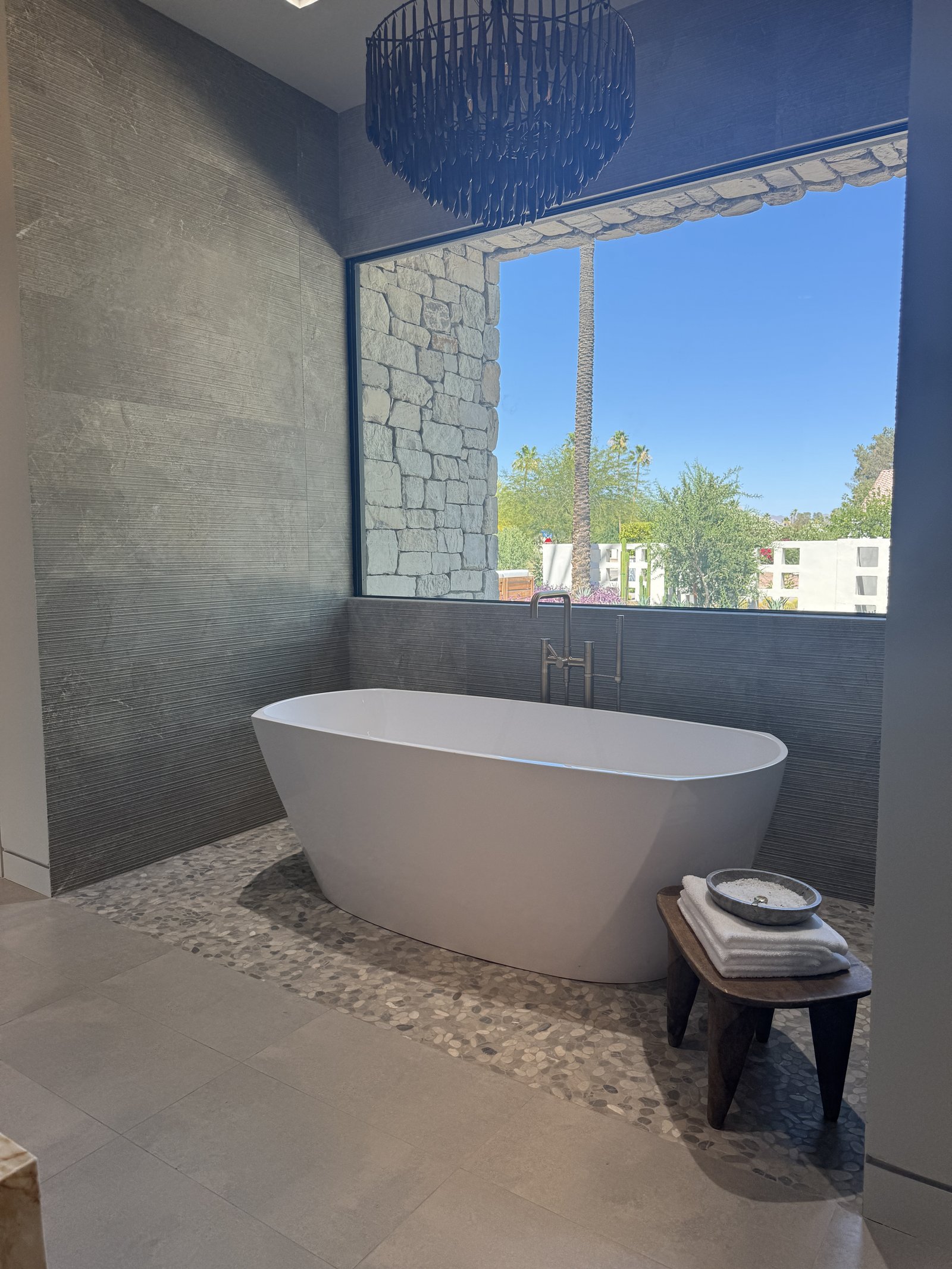 Gilbert AZ primary bathroom with freestanding tub on pebble-finish floor tile, hand-beaded blue chandelier, and a landscape-view picture window