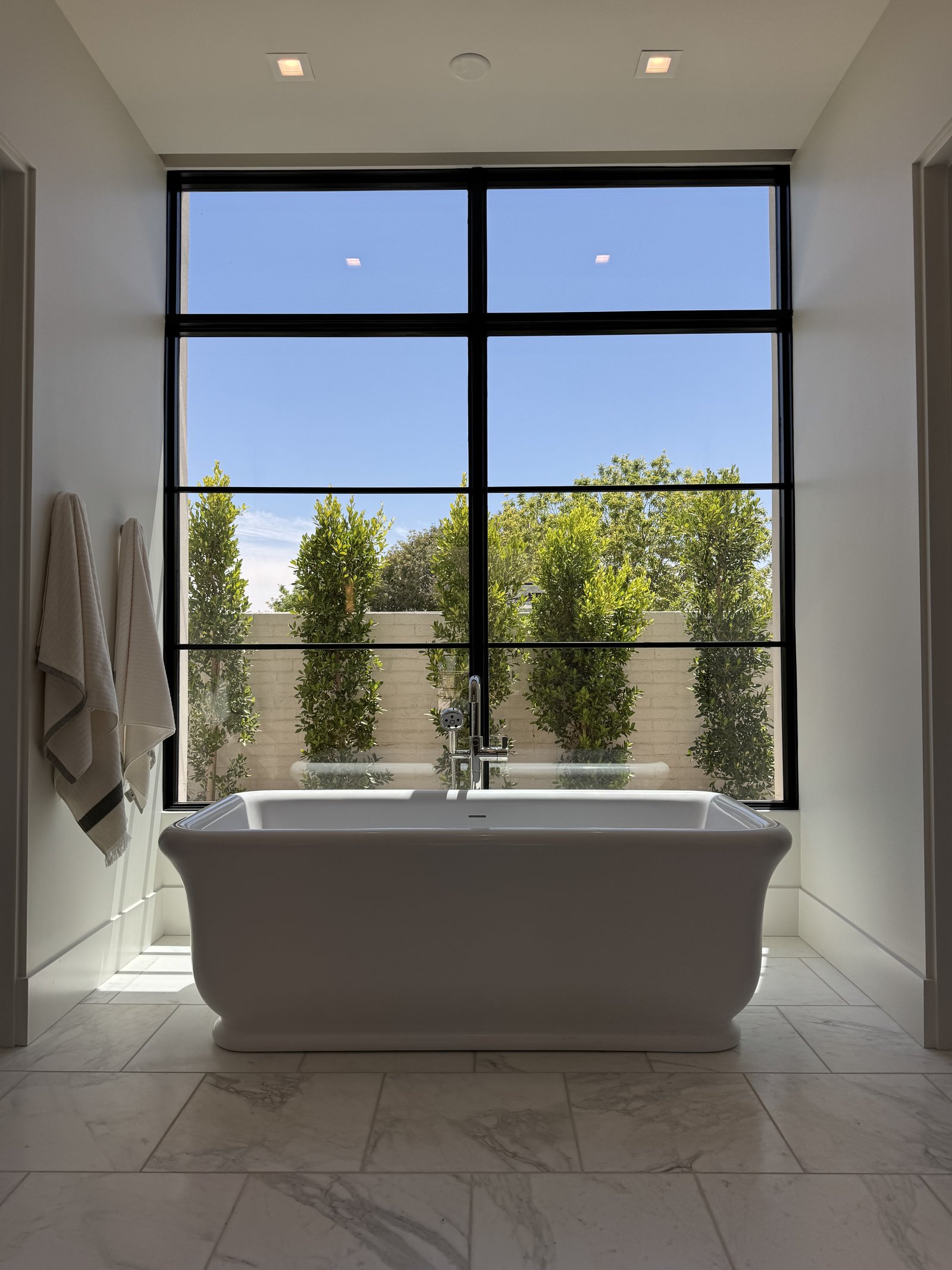 Phoenix AZ primary bathroom remodel with sculptural freestanding tub, floor-to-ceiling black-grid window, and herringbone marble floor