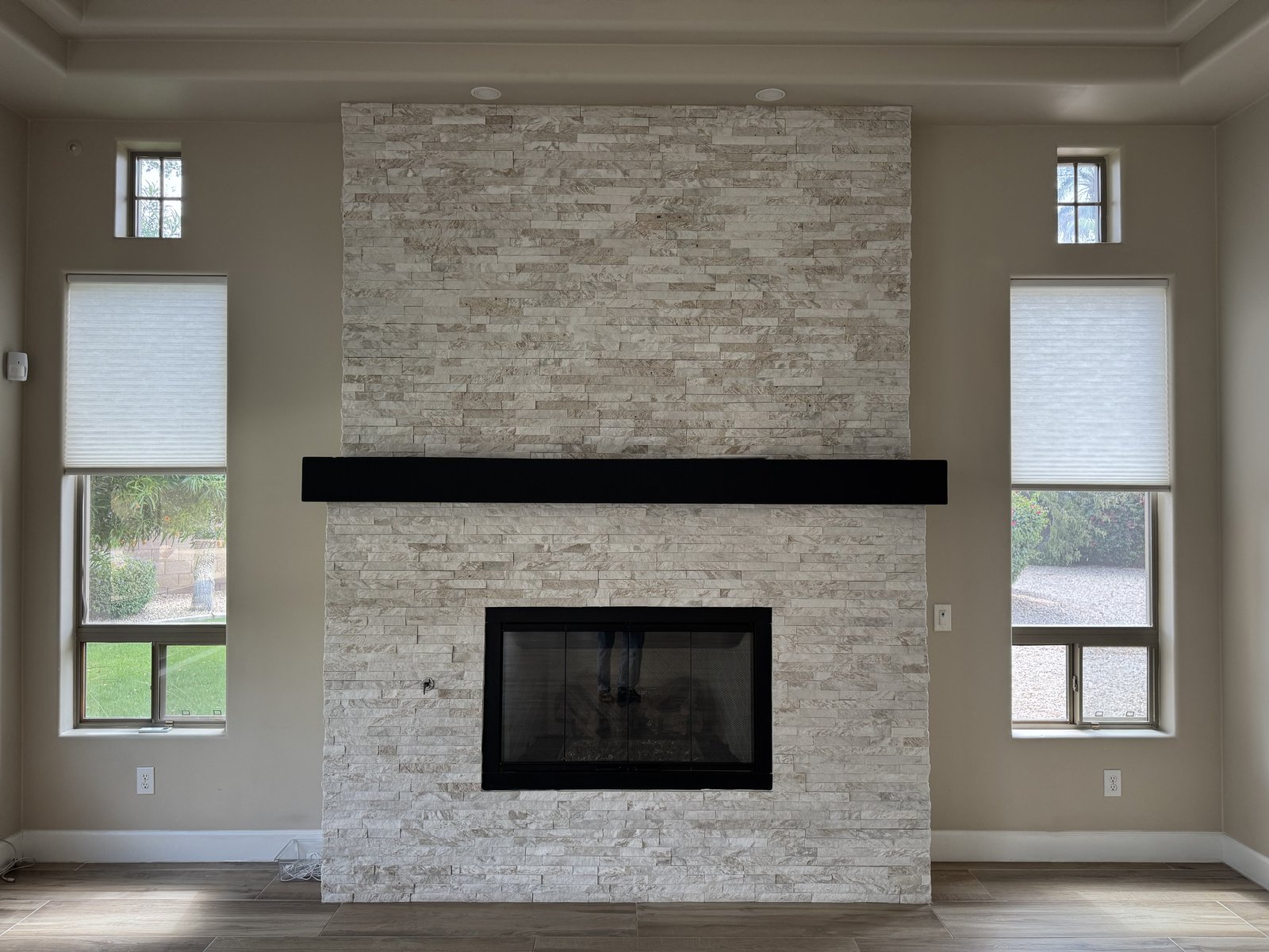 Queen Creek AZ custom home stacked-stone fireplace feature wall with solid dark-wood mantel and flanking trim-set windows, shown near finish phase